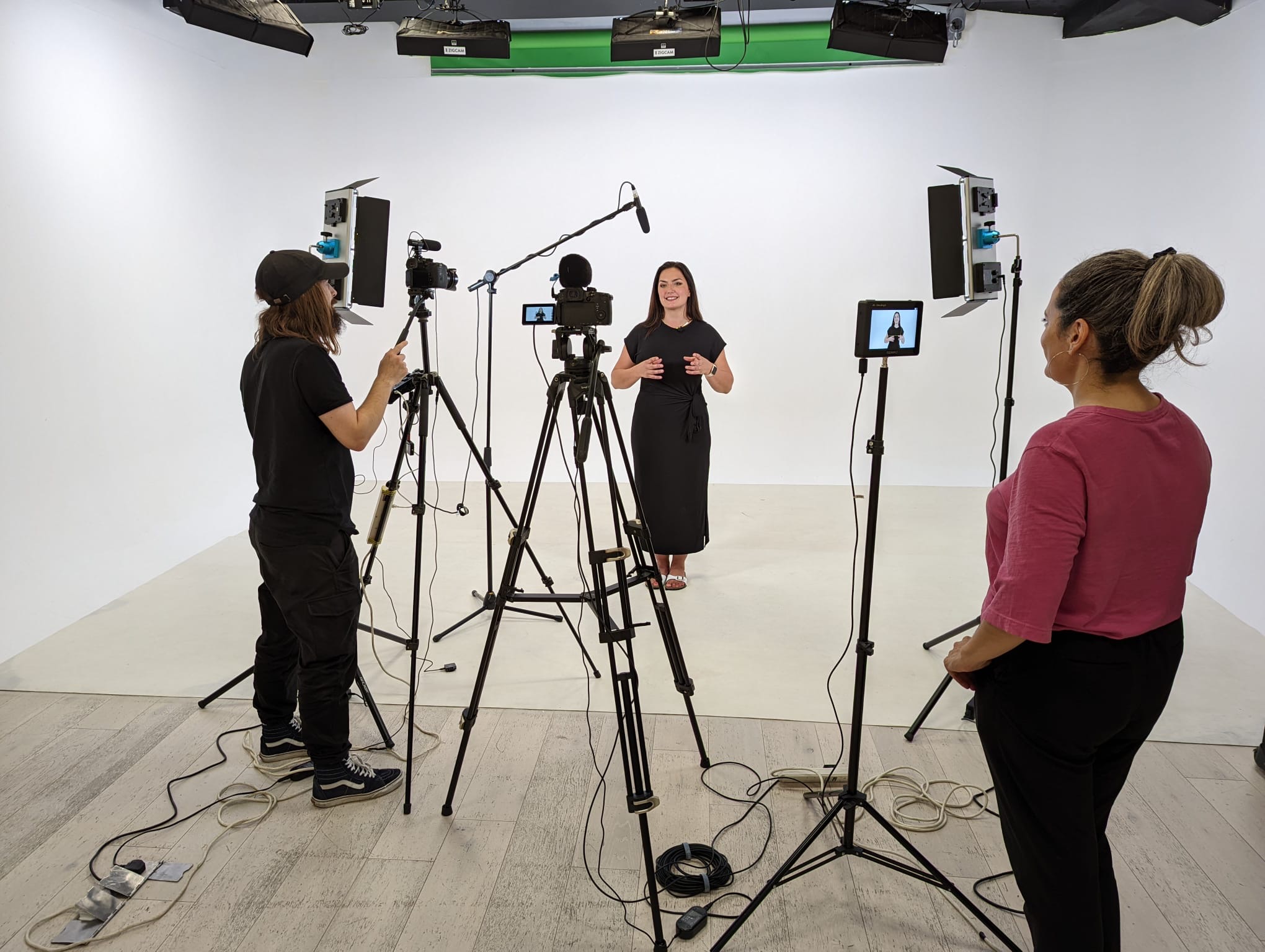 A small filming set-up in a studio, against a white screen. Emma Hill is looking at the monitor. Anna Patalong is being filmed and Chris Forshaw is operating the camera.